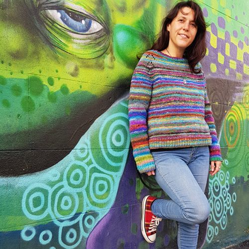 standing woman leaning against a green graffitti wall wearing a hand knit sweater felix pullover designed by amy christoffers in colorful self striping yarn noro ito